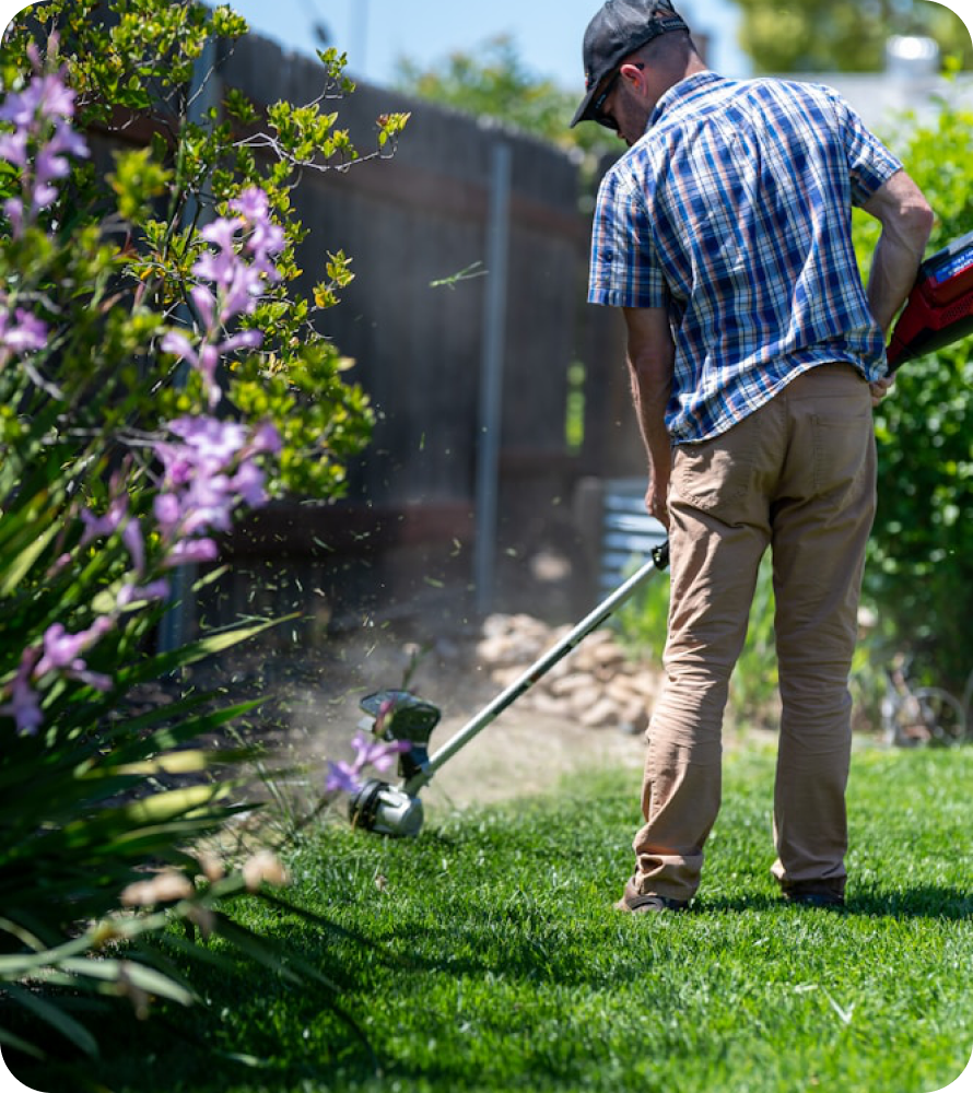 A Soils Alive technician using a string trimmer along a flower bed at the edge of a lawn