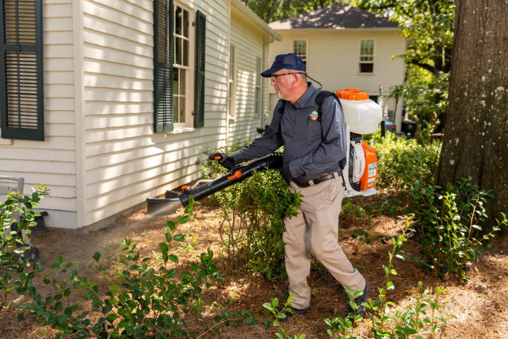 A technician with a backpack mist blower treating shrubs beside a house